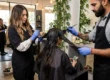 Deux coiffeurs travaillent sur une cliente, appliquant une coloration naturelle des cheveux avec des outils et gants bleus.